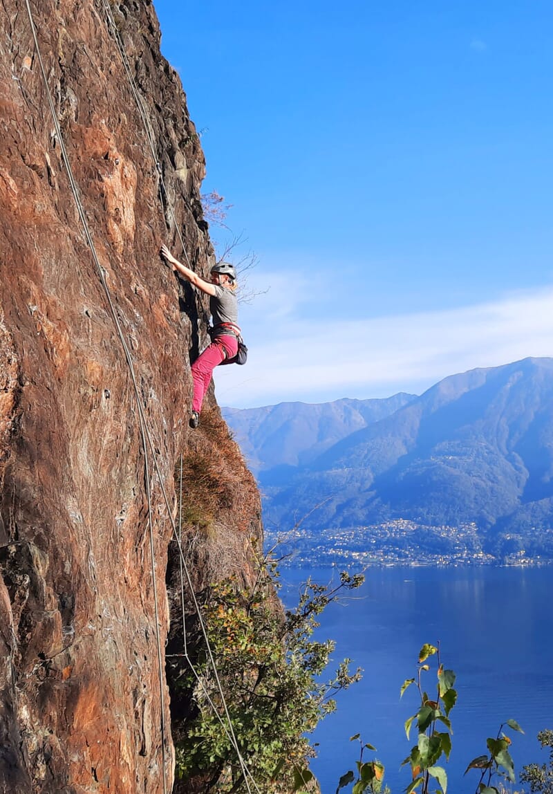 Kletterin im Nachstieg in Felswand mit Lago Maggiore im Hintergrund